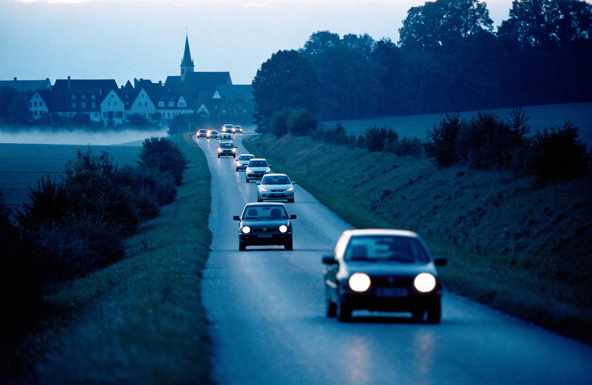 Berufspendler auf einer Landstraße im Morgengrauen Autoschlange auf einer Landstraße im Morgengrauen vor einem Dorf – KI-generiertes Symbolbild